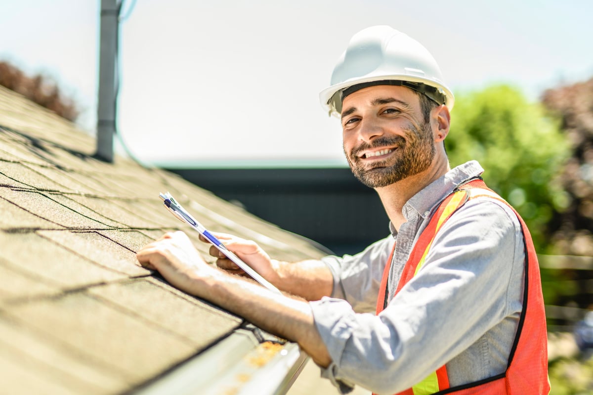 Roofing inspector with a white hard hat holding a clipboard inspecting a house